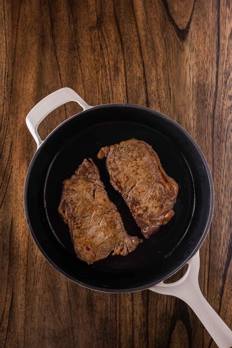 Two cooked steaks in a black cast iron skillet with a white handle, placed on a wooden surface.