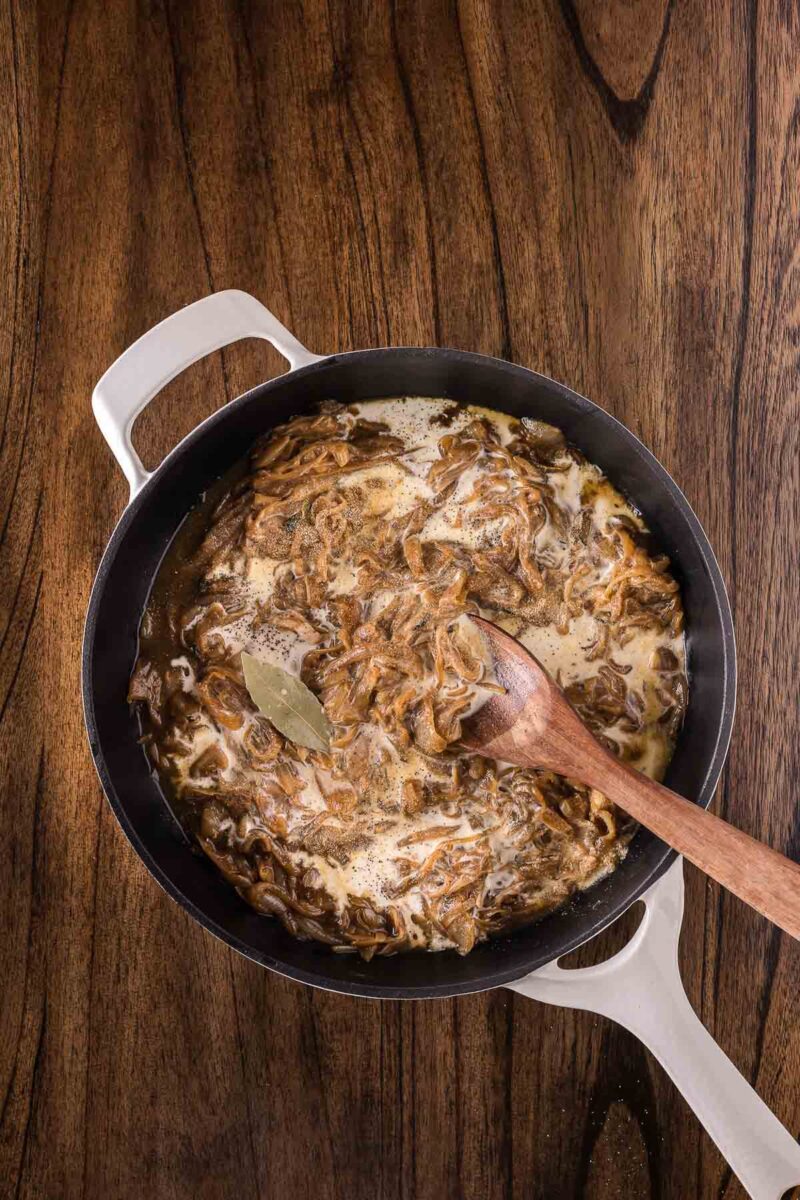 A white skillet on a wooden surface contains caramelized onions in a creamy sauce with a bay leaf; a wooden spoon rests inside the pot.
