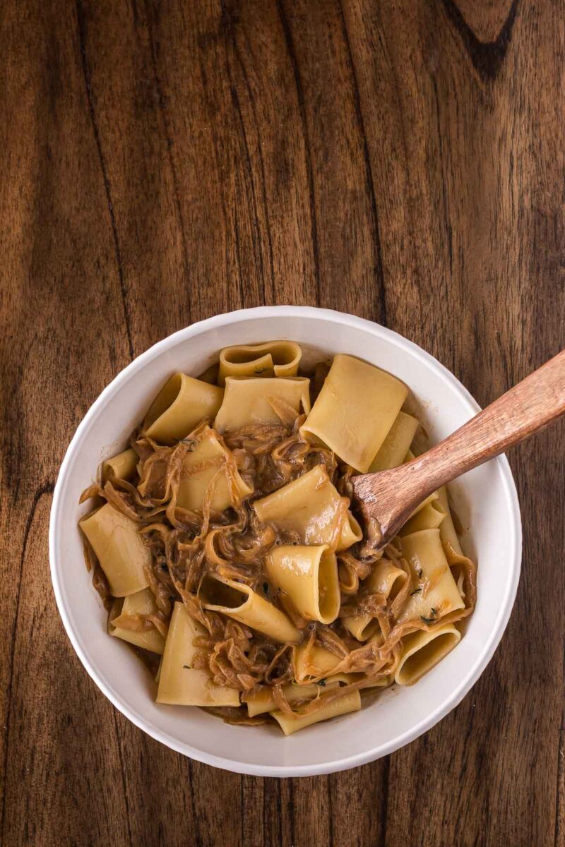 A white bowl filled with wide pasta noodles in a creamy brown onion sauce, with a wooden spoon resting inside, placed on a wooden table.