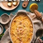 Baked 4 cheese hot lobster dip in an oval dish, surrounded by seasoning bowls, herbs, lemon, and serving utensils on a rustic surface.