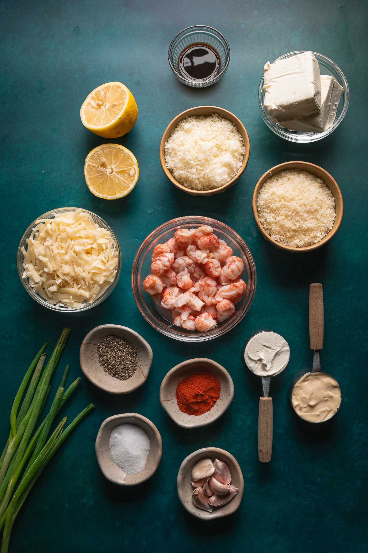 Top-down view of various ingredients in bowls and dishes, including cooked lobster, cheeses, seasonings, lemon halves, green onions, and creamy sauces on a dark green surface.