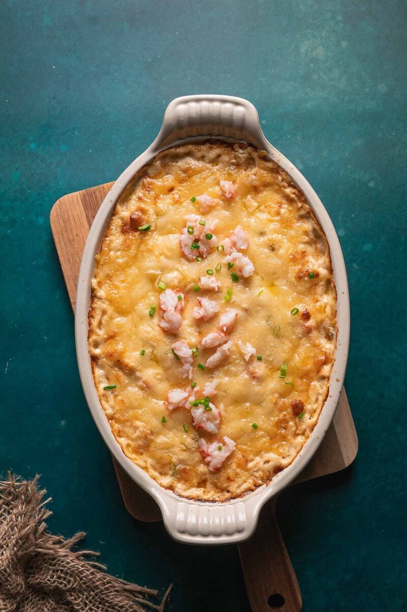 Oval baking dish filled with cheesy hot lobster dip topped with chopped chives, placed on a wooden board against a blue background.