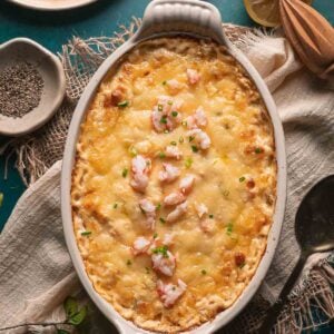 Oval casserole dish filled with baked hot lobster dip topped with chives; surrounded by serving utensils, pepper, and half a lemon.