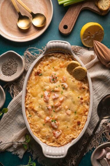 A baked cheesy hot lobster dip, garnished with herbs and served with two round toasted bread pieces in a white oval dish.