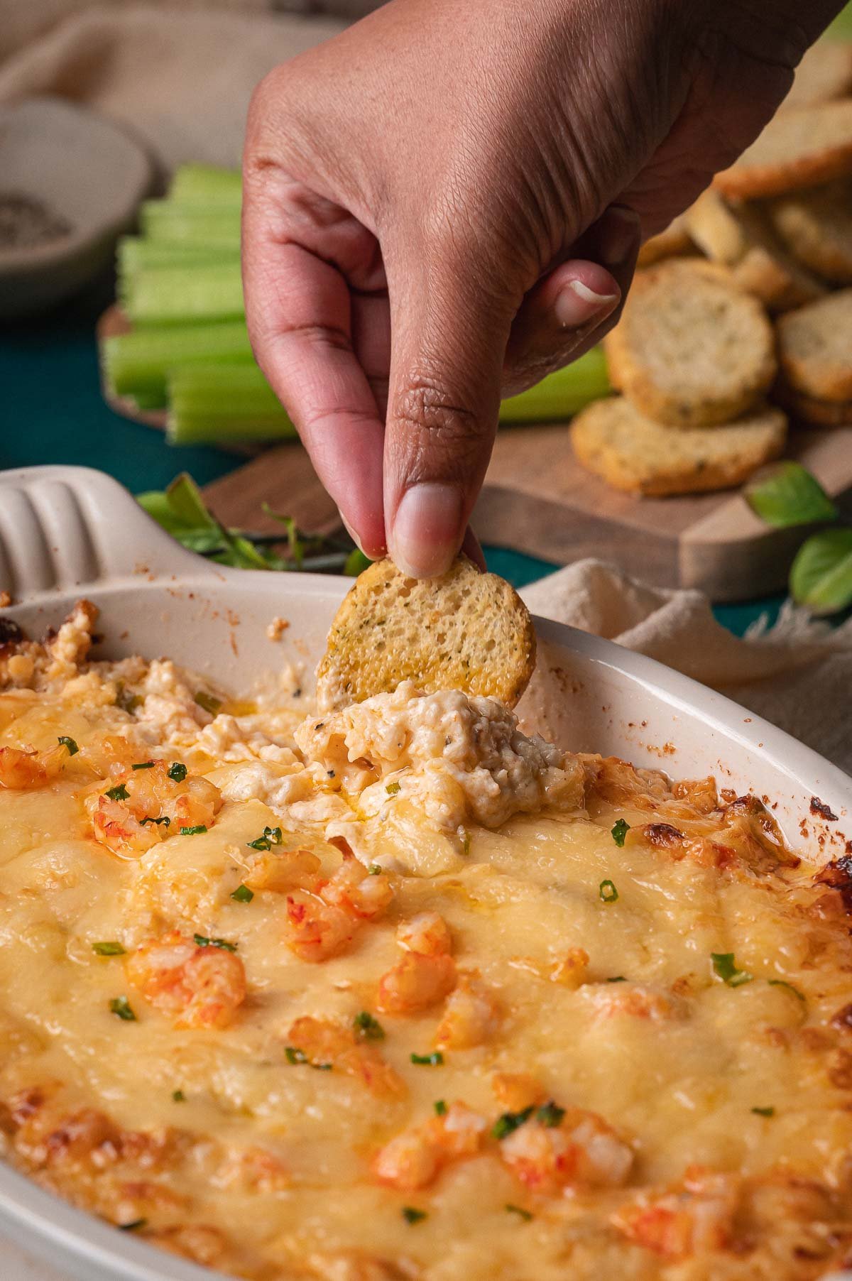 A hand dips a toasted bread round into a cheesy baked hot lobster dip dish topped with herbs, with celery and more bread in the background.