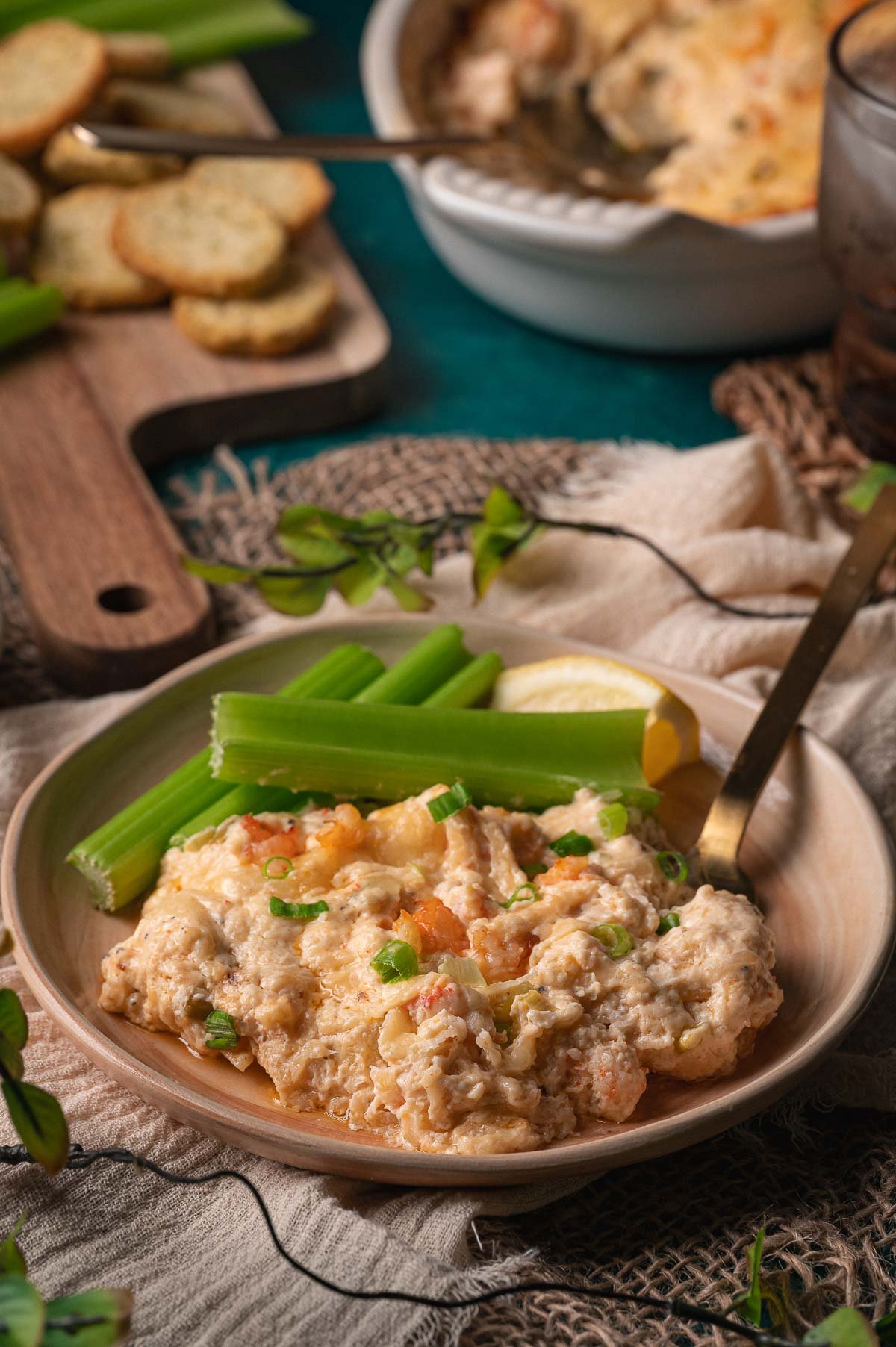 A plate with creamy hot lobster dip garnished with green onions, served alongside celery sticks and a lemon wedge, with crackers and more dip in the background.