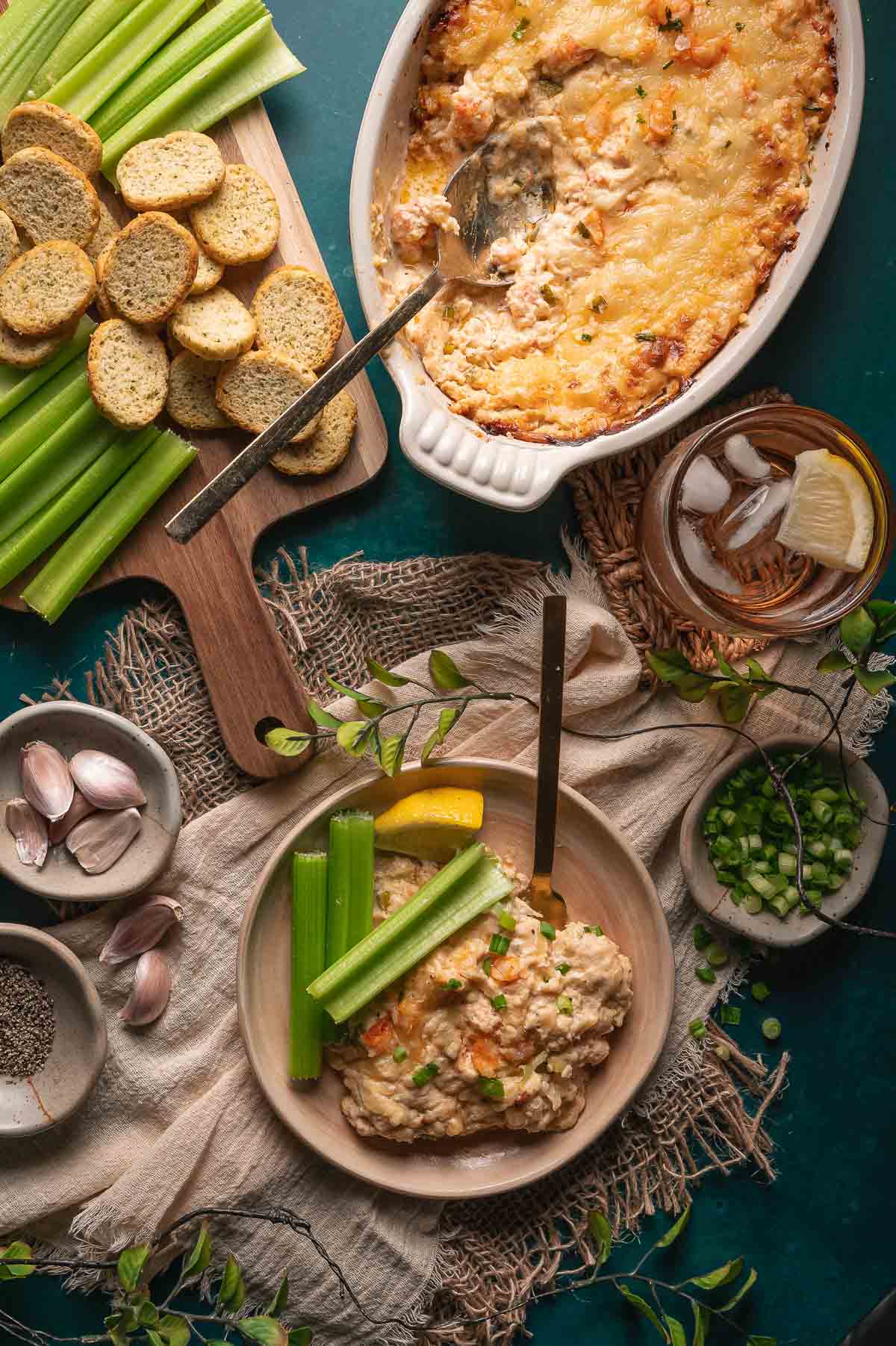 A bowl of creamy hot lobster dip with celery sticks and a lemon wedge, next to a baked dish of dip, toasted bread slices, a glass of iced drink, sliced green onions, garlic, and pepper.