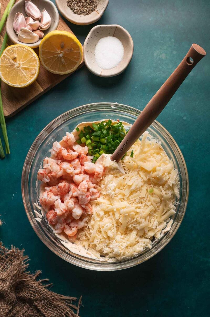 A glass bowl contains shredded cheese, chopped green onions, and pieces of lobster. Lemon, salt, pepper, garlic, and chives are arranged nearby on the table.