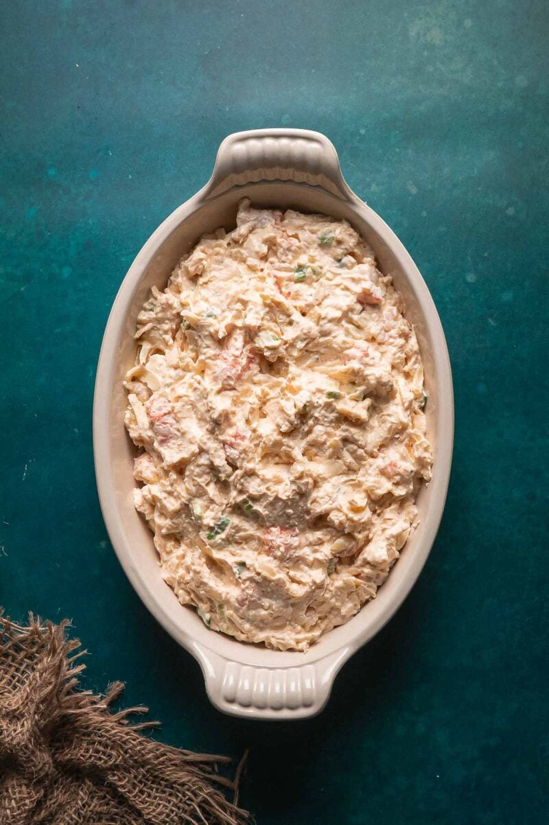 Oval dish filled with a creamy mixture of lobster dip containing visible bits of lobster meat, set on a dark teal background with a piece of burlap fabric nearby.