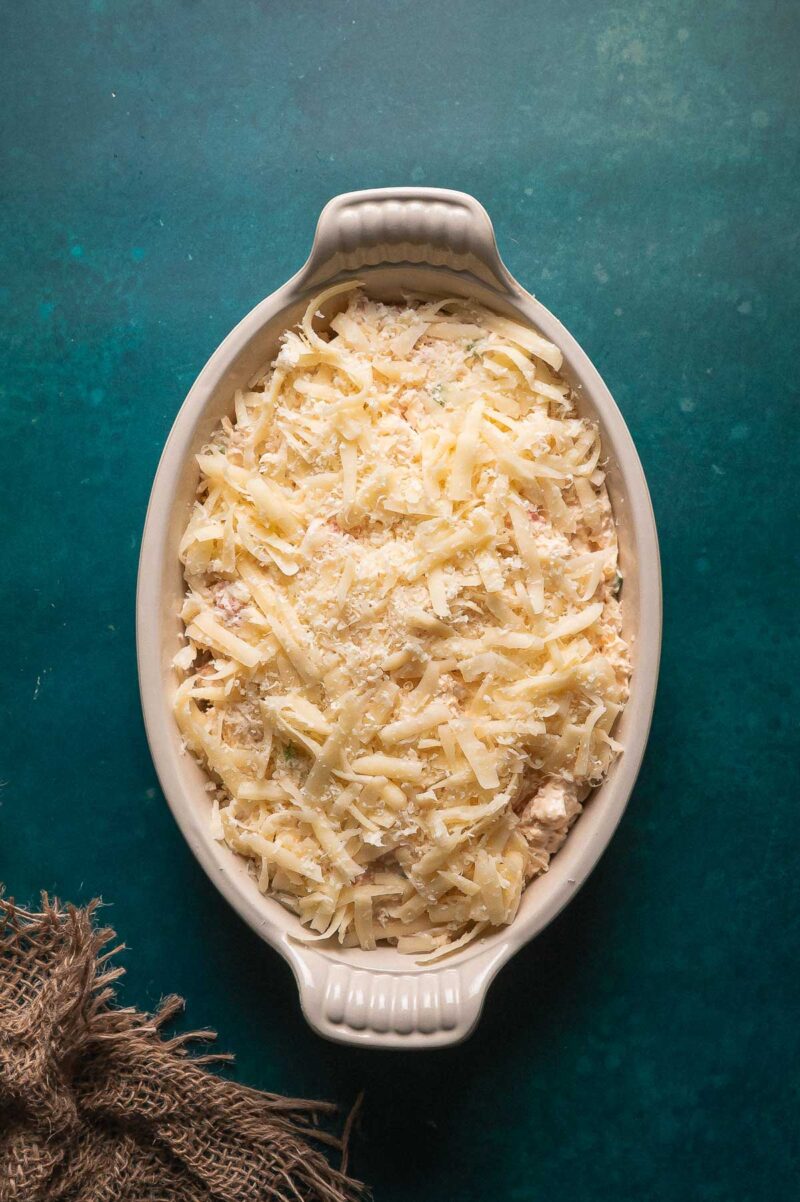 An oval baking dish filled with an unbaked hot lobster dip topped with shredded cheese on a teal surface.