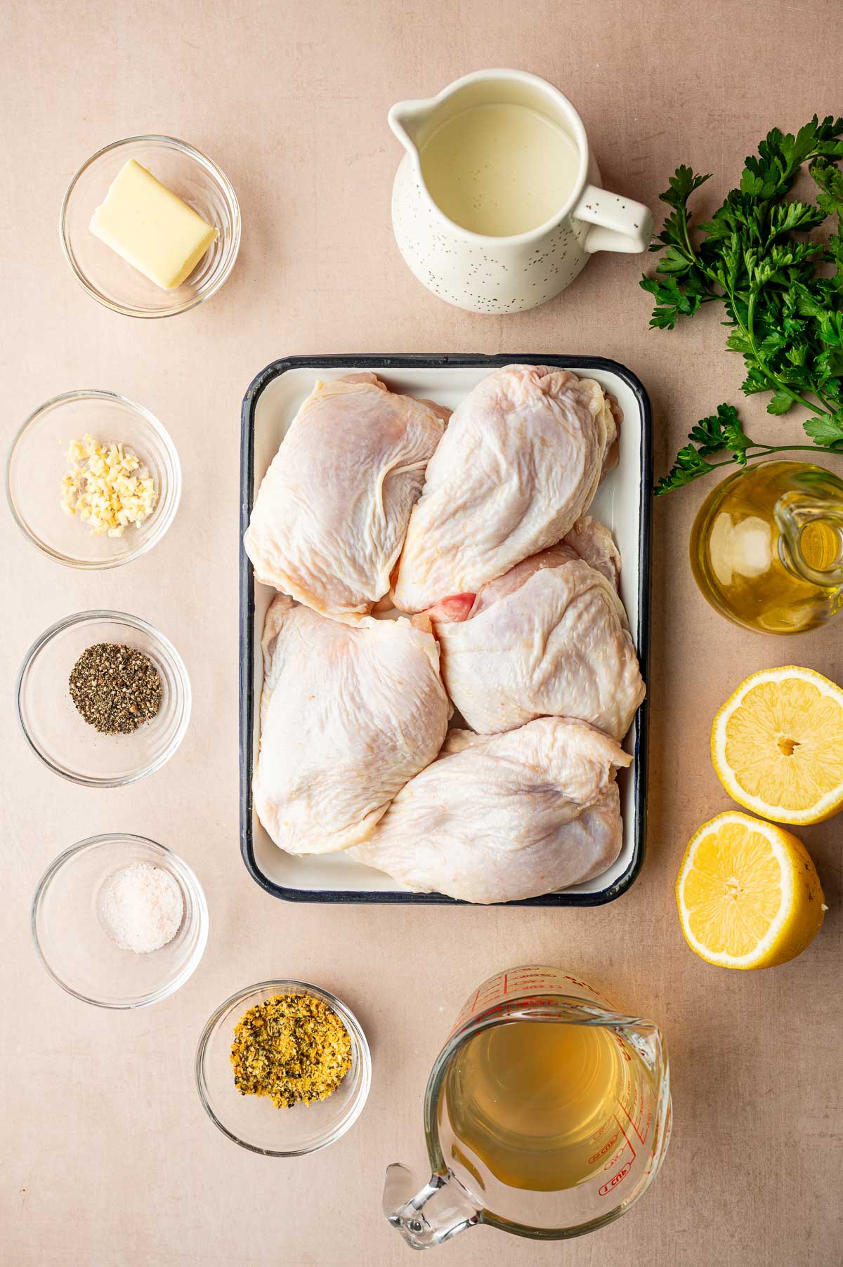 Raw chicken thighs in a tray surrounded by butter, minced garlic, salt, pepper, seasoning, lemon, olive oil, broth, and parsley arranged on a light tabletop.