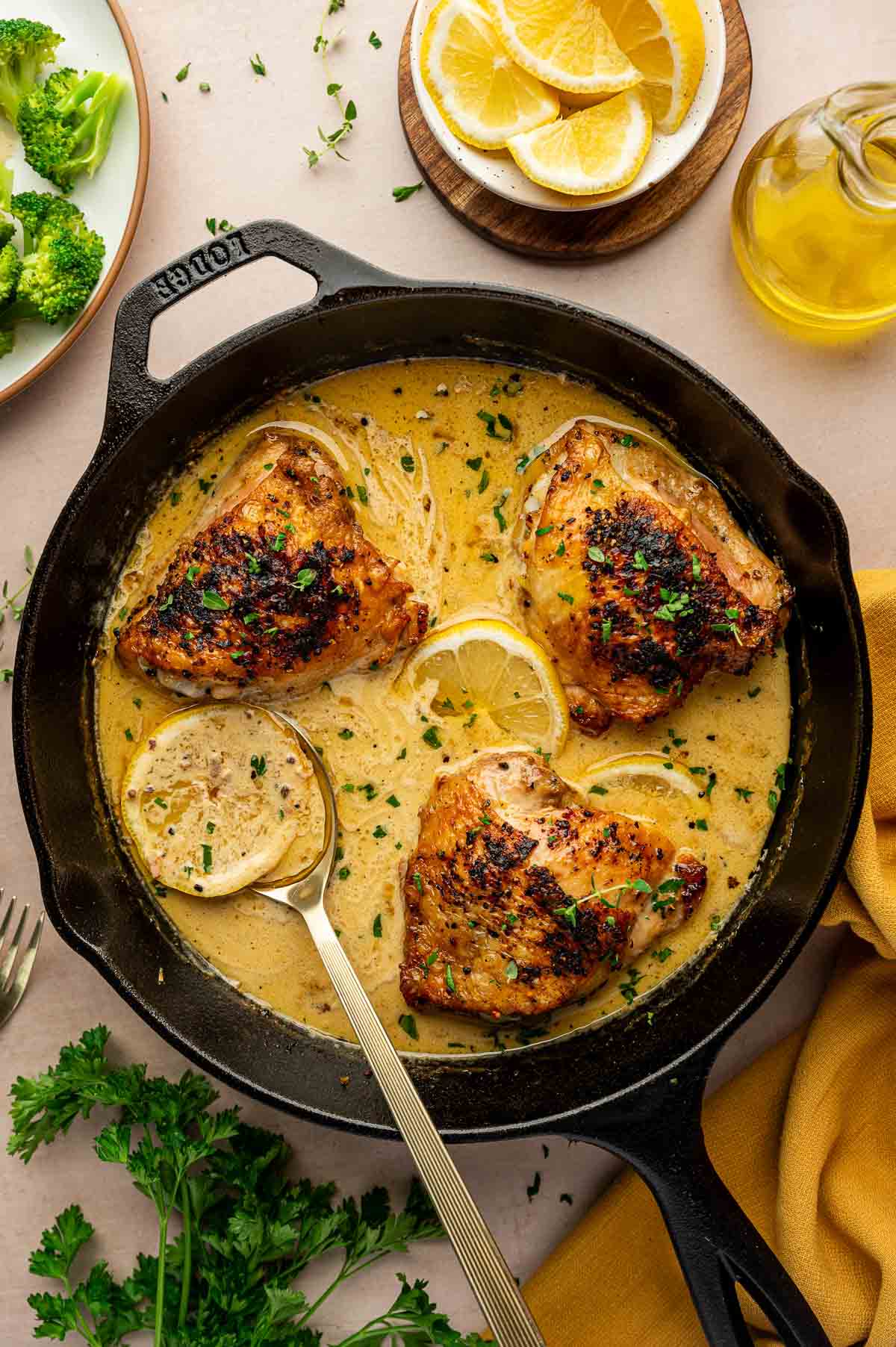 Three cooked chicken thighs in a creamy lemon pepper sauce with lemon slices in a cast iron skillet, garnished with herbs; a serving spoon rests in the pan. Lemons, broccoli, and olive oil nearby.