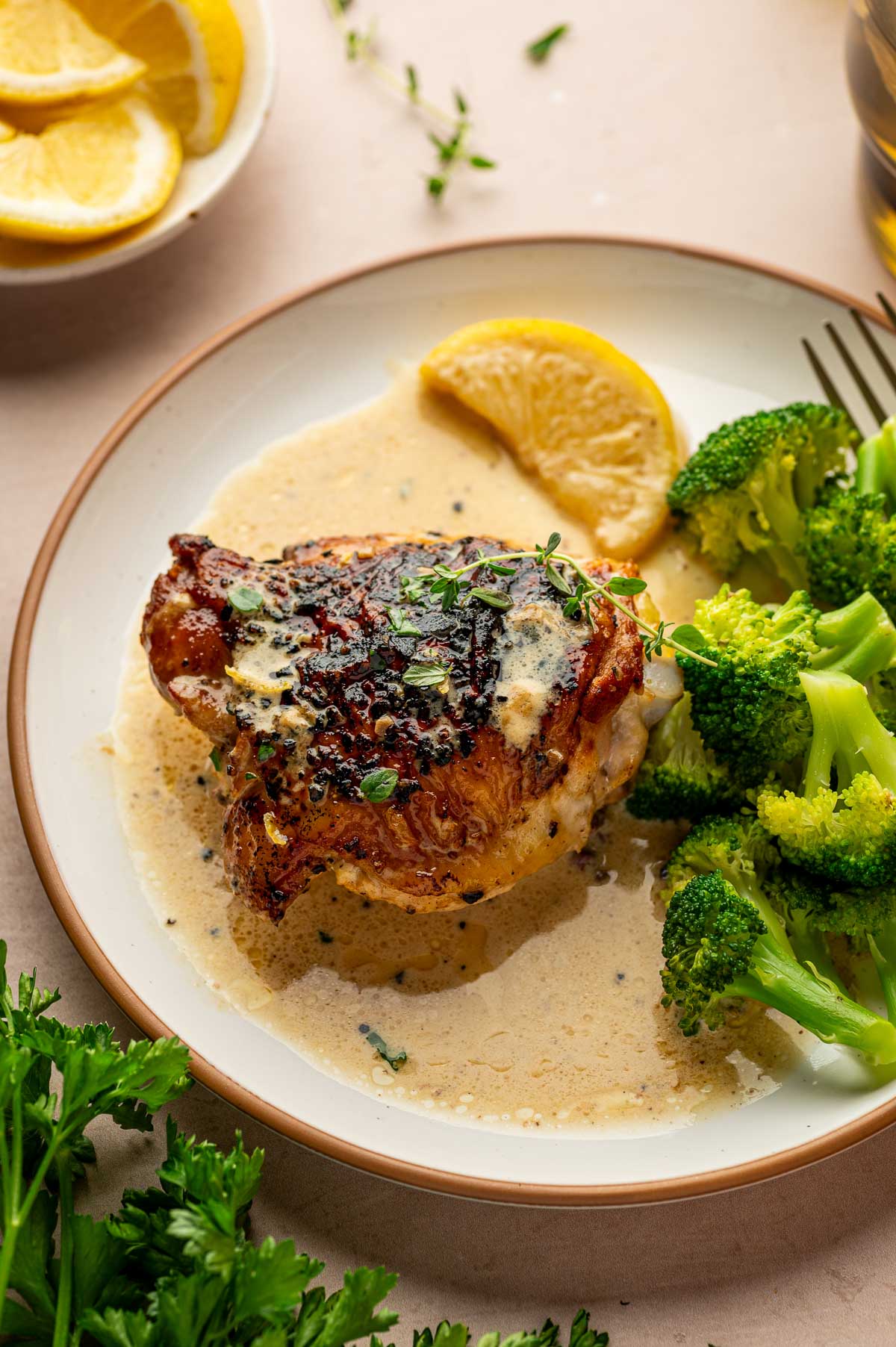 A plate with a chicken thigh n in a creamy lemon pepper sauce, garnished with herbs, served with steamed broccoli and a lemon wedge.