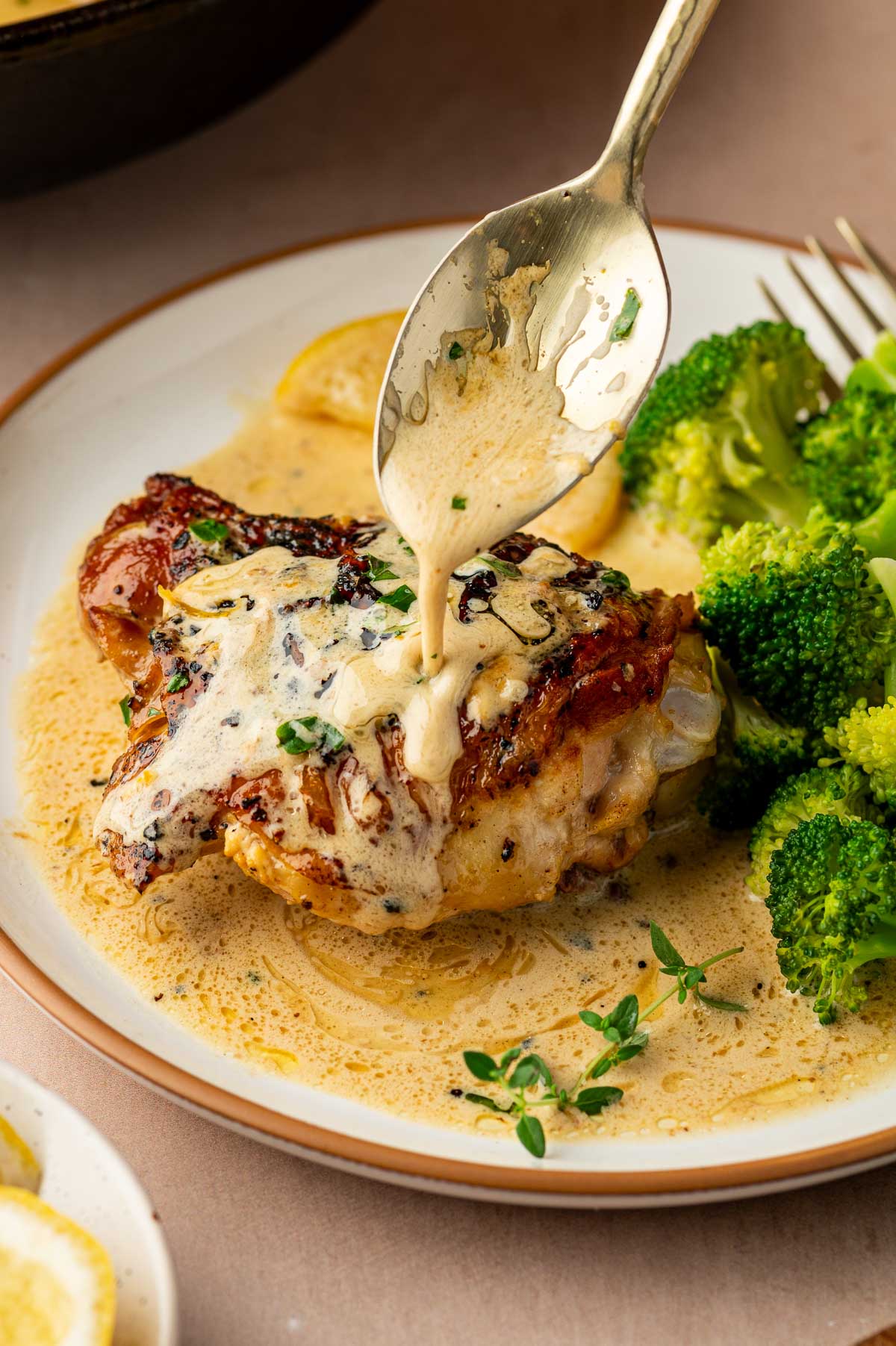 A spoon pours creamy lemon pepper sauce over a cooked chicken thigh on a plate with steamed broccoli and fresh herbs.