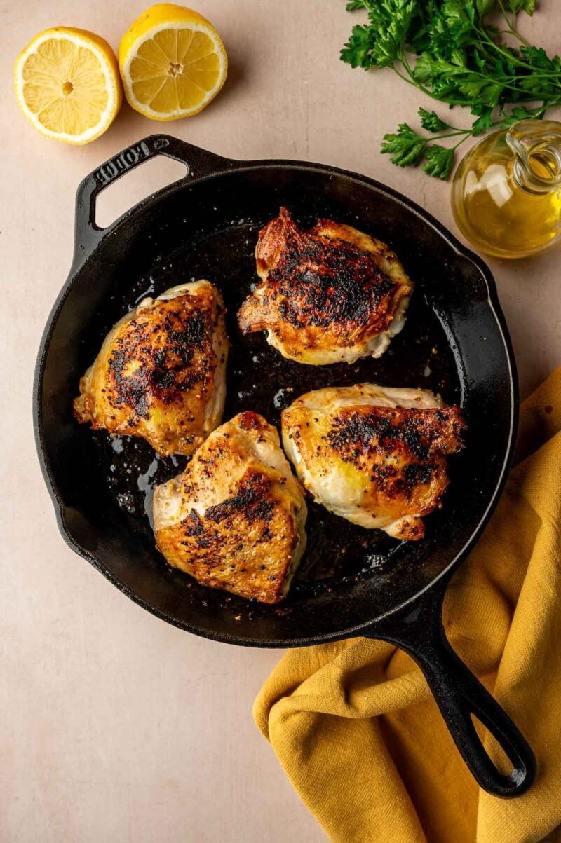 Four seasoned, cooked chicken thighs in a cast iron skillet, with a yellow cloth, lemon halves, fresh parsley, and a bottle of olive oil nearby.