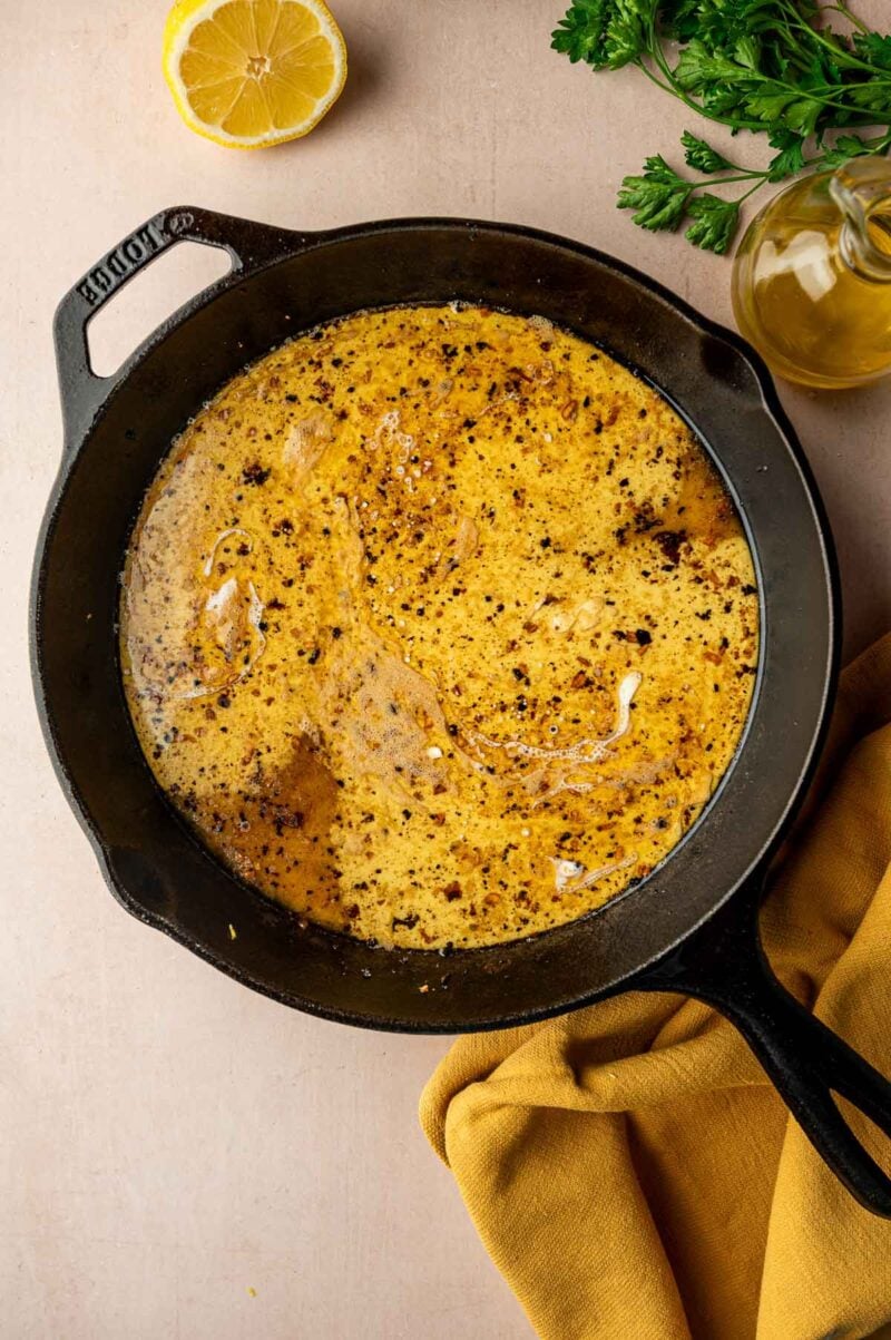 A cast iron skillet filled with a seasoned, creamy sauce sits on a beige surface near a lemon half, fresh herbs, a bottle of oil, and a yellow cloth.