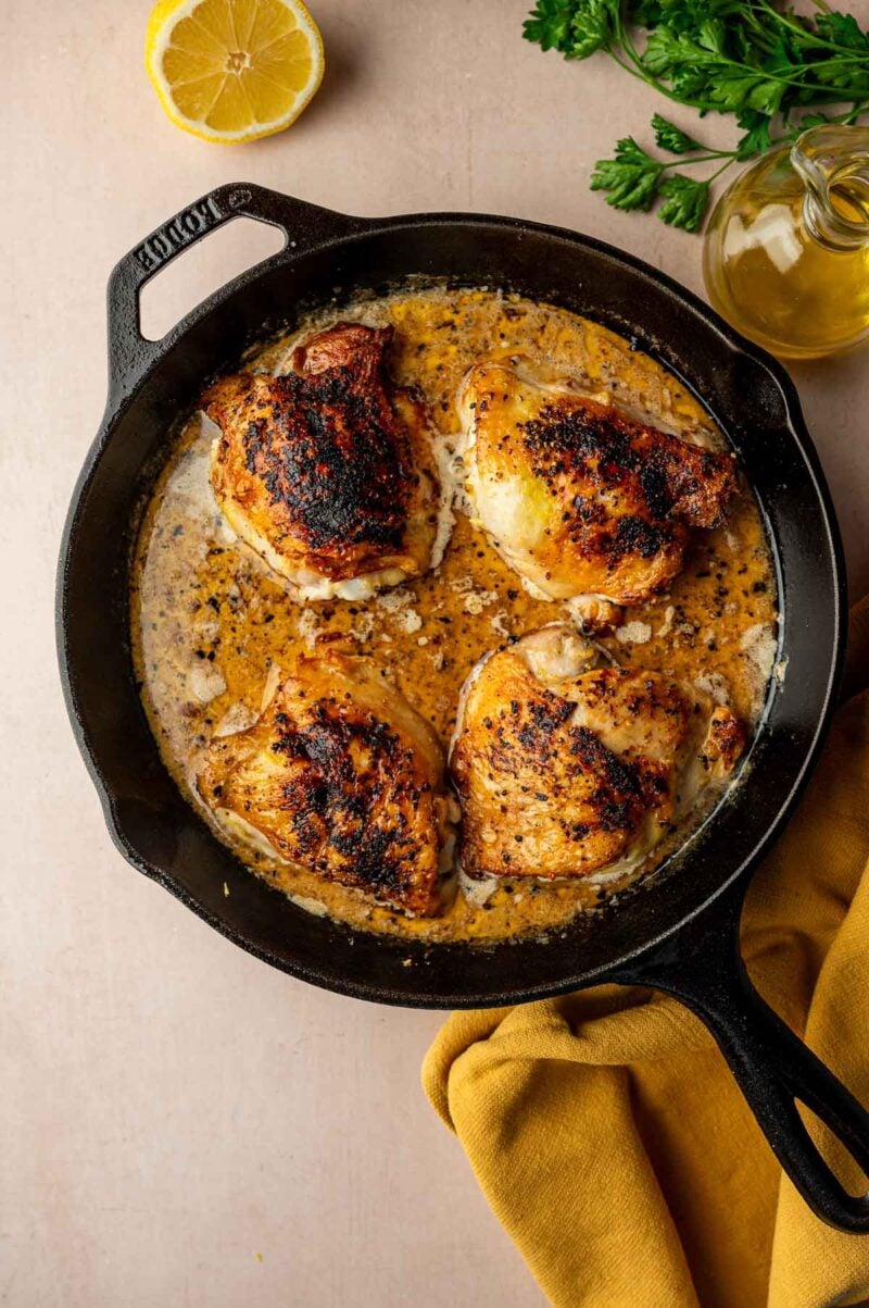 A cast iron skillet containing four seasoned, cooked chicken thighs in lemon pepper sauce, surrounded by a lemon half, parsley, a bottle of oil, and a yellow cloth.