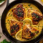 Four browned chicken thighs in a creamy lemon pepper sauce with lemon slices and herbs, cooked in a cast iron skillet, surrounded by broccoli, parsley, lemon wedges, and olive oil.