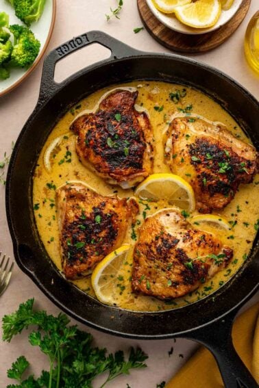 Four browned chicken thighs in a creamy lemon pepper sauce with lemon slices and herbs, cooked in a cast iron skillet, surrounded by broccoli, parsley, lemon wedges, and olive oil.