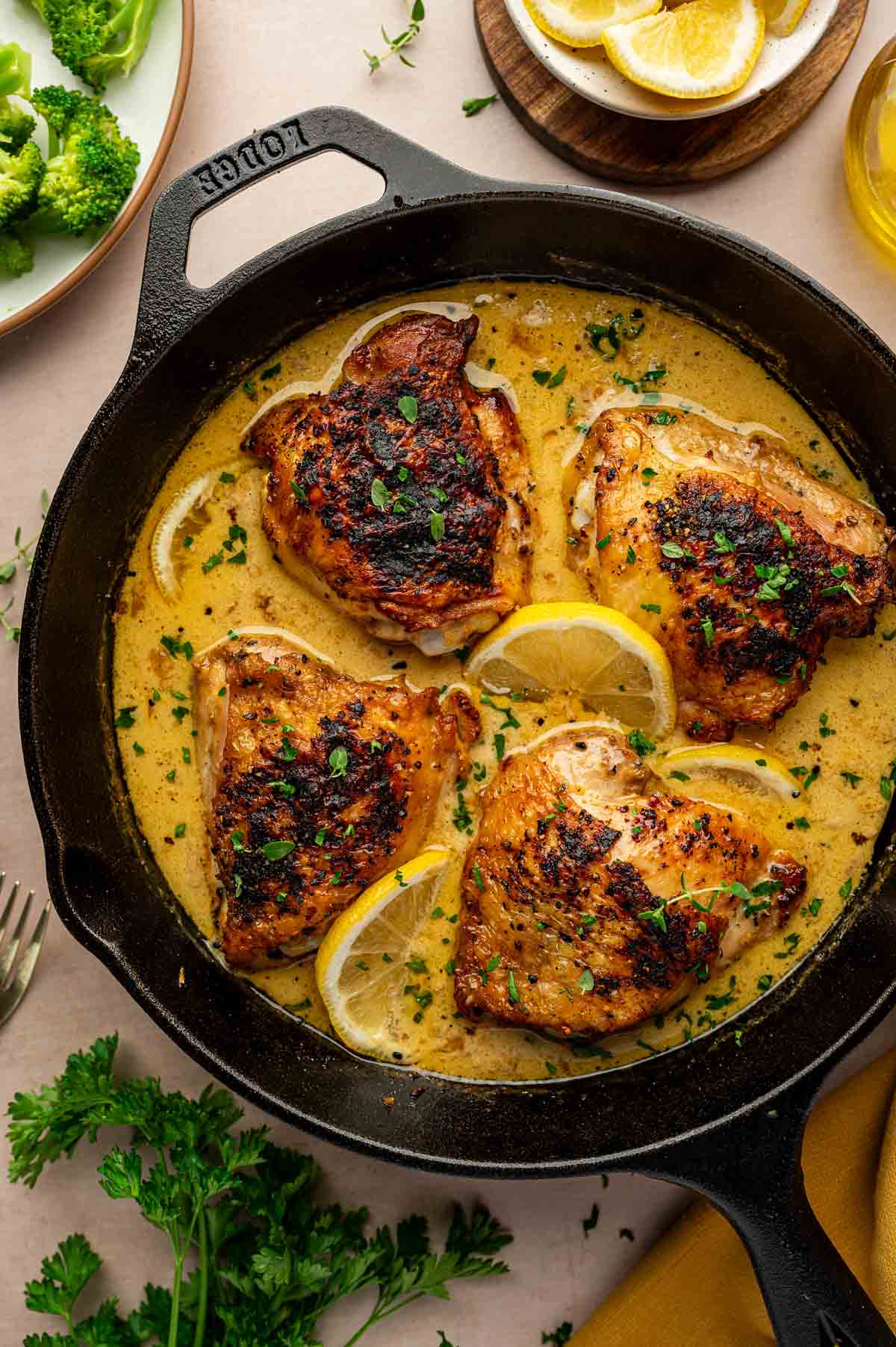 Four browned chicken thighs in a creamy lemon pepper sauce with lemon slices and herbs, cooked in a cast iron skillet, surrounded by broccoli, parsley, lemon wedges, and olive oil.