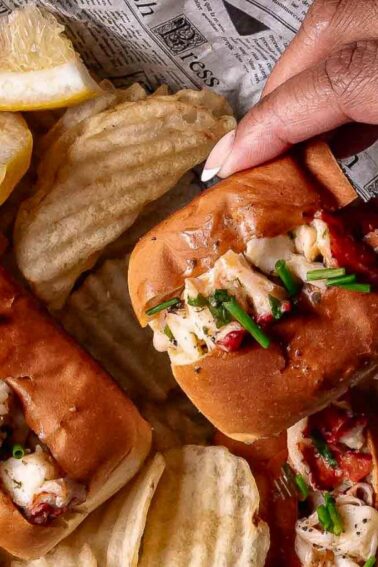 A hand picking up a lobster roll from a plate with lobster rolls, potato chips, and lemon wedges on newspaper-like paper.