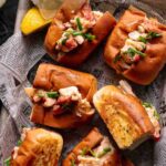 Overhead view of Connecticut style lobster rolls on toasted buns, garnished with chives and served with lemon wedges on newspaper. Text overlay includes recipe source information.