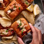 A hand holds a lobster roll topped with chives, surrounded by more lobster rolls, potato chips, and lemon wedges on newspaper-lined tray.
