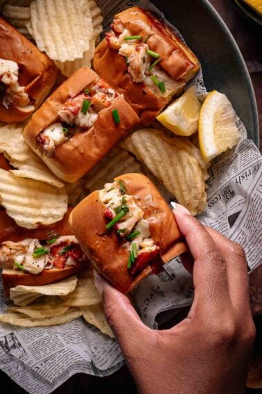A hand holds a lobster roll topped with chives, surrounded by more lobster rolls, potato chips, and lemon wedges on newspaper-lined tray.