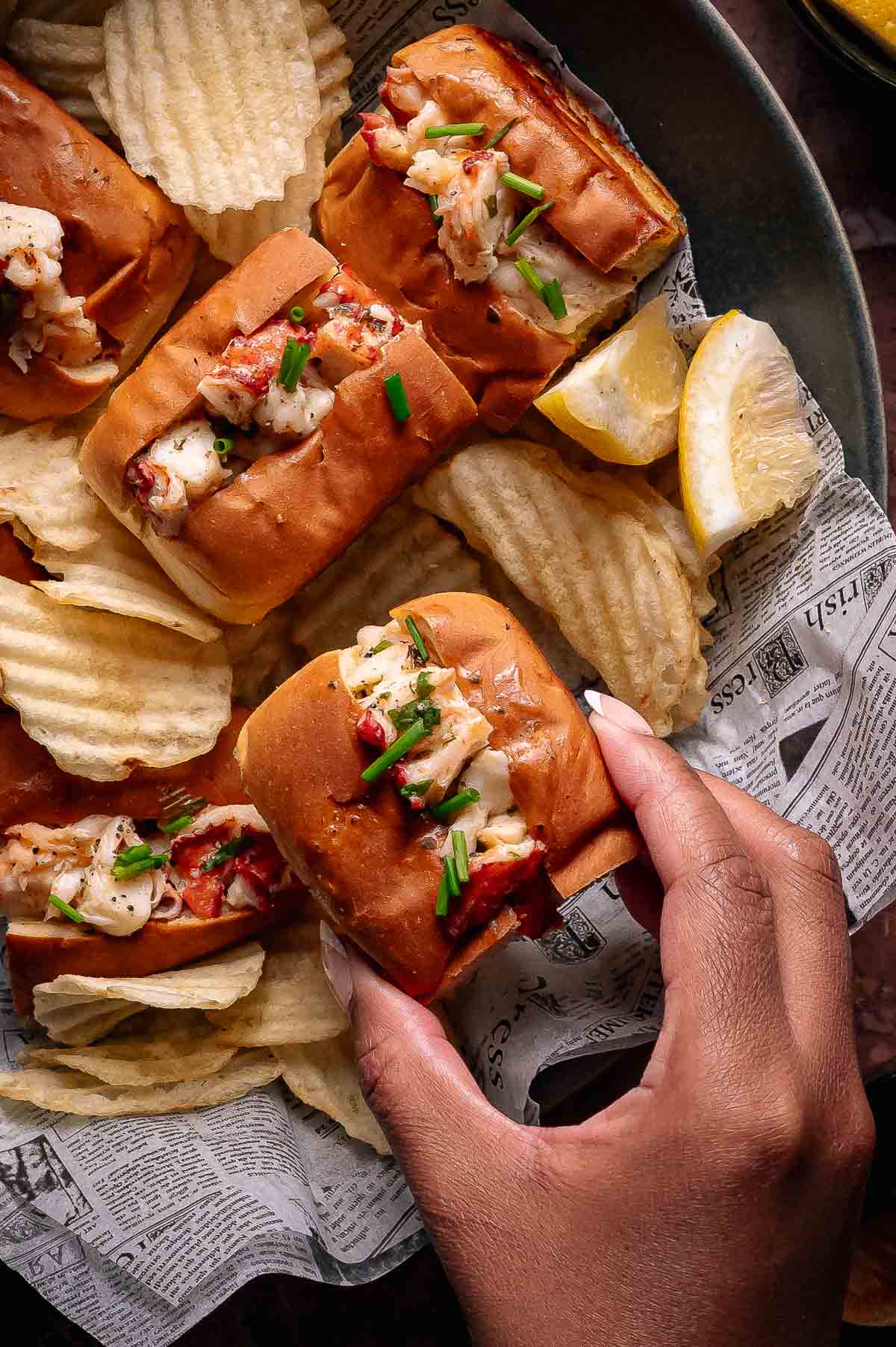 A hand holds a lobster roll topped with chives, surrounded by more lobster rolls, potato chips, and lemon wedges on newspaper-lined tray.