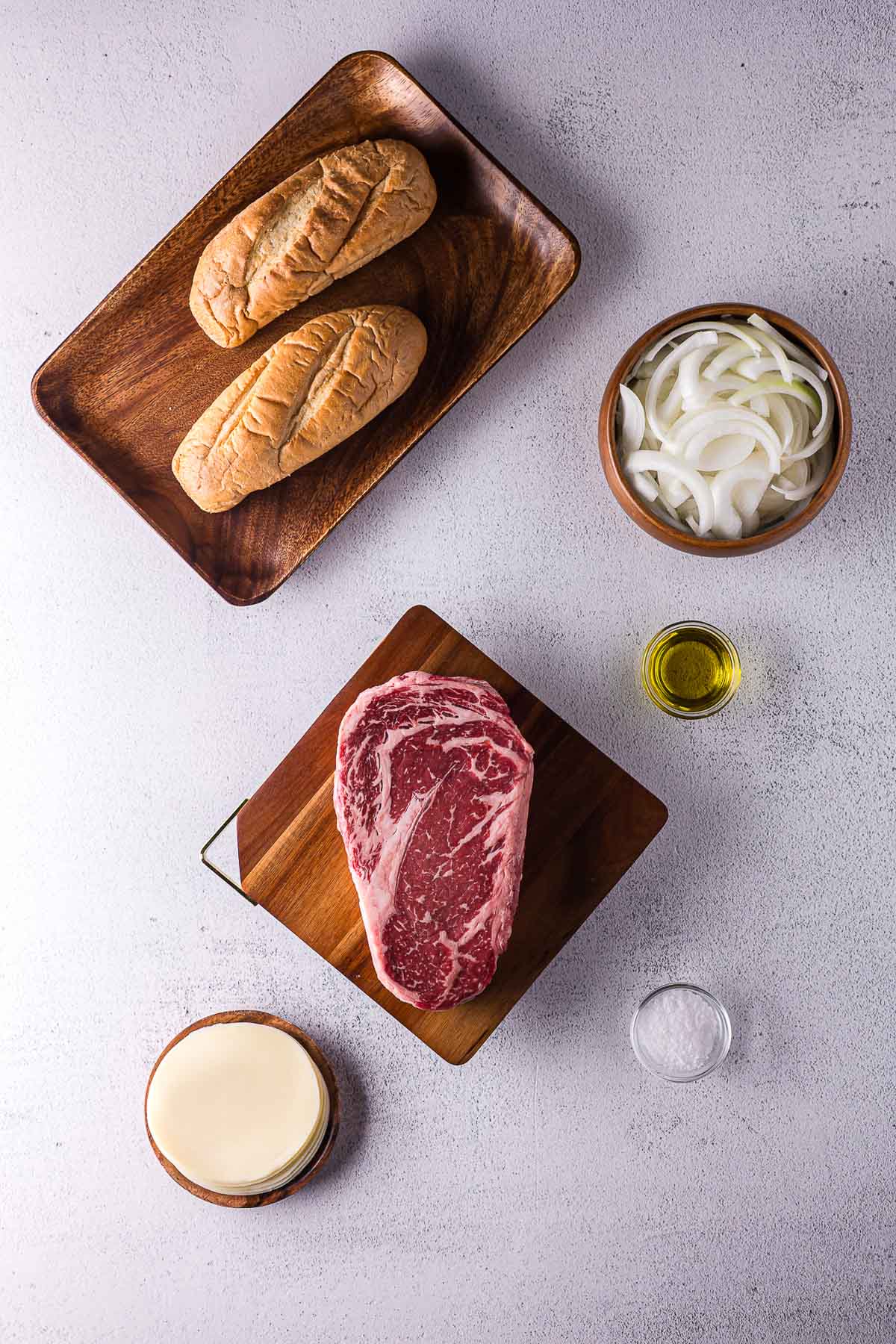 A steak, two bread rolls, sliced onions, a small bowl of olive oil, a bowl of salt, and sliced cheese are arranged on a white surface.