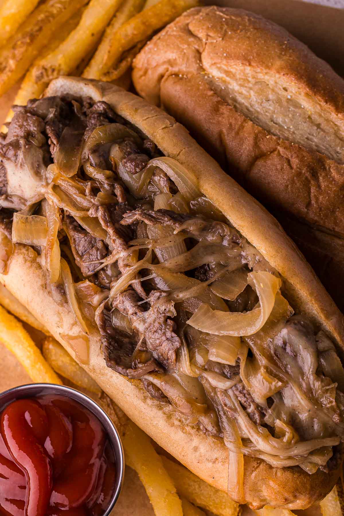 A Philly cheesesteak sandwich with sautéed onions, served with French fries, a side of ketchup, and a hoagie roll.
