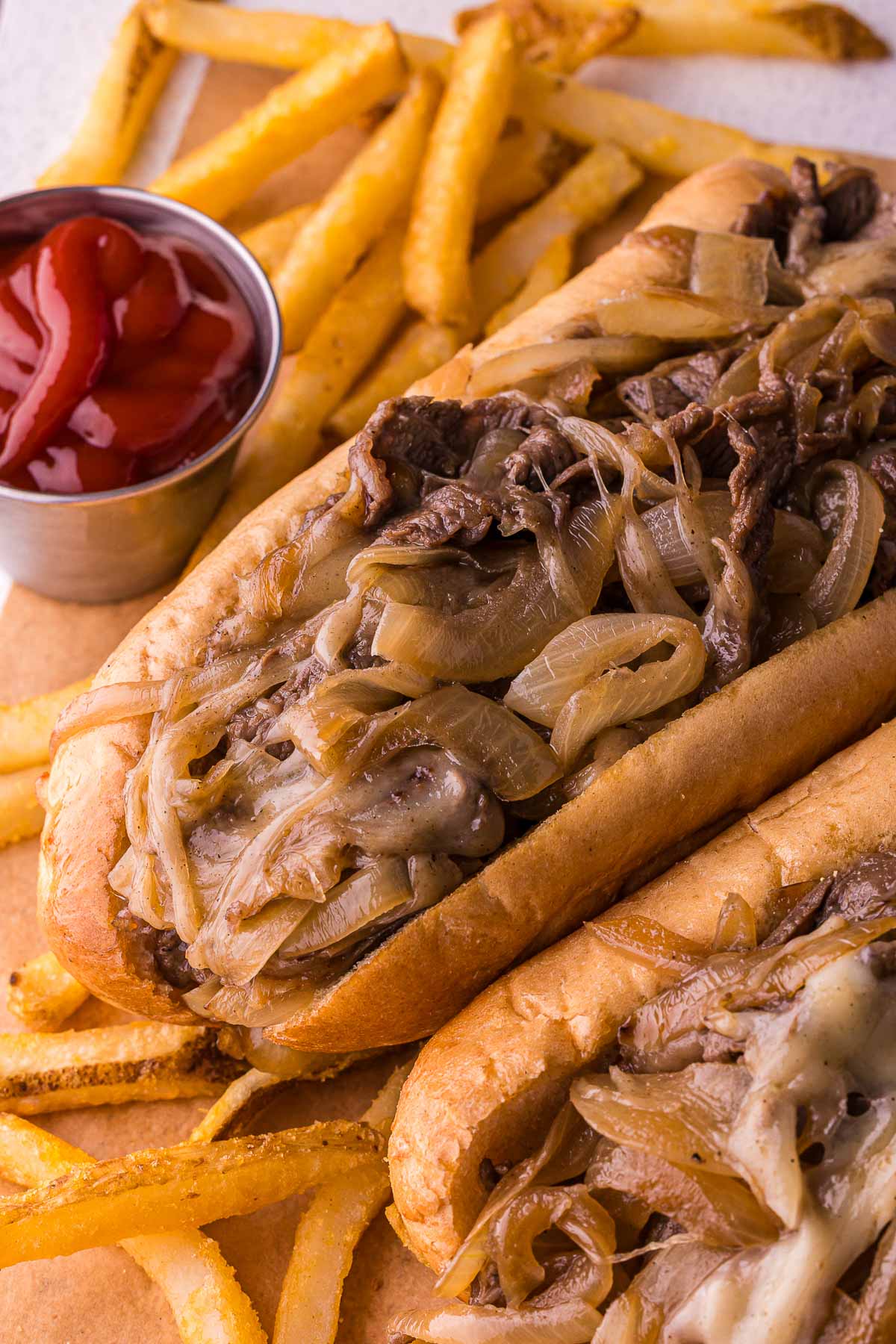 A close-up of two cheesesteaks with onions and a side of French fries and a small cup of ketchup.