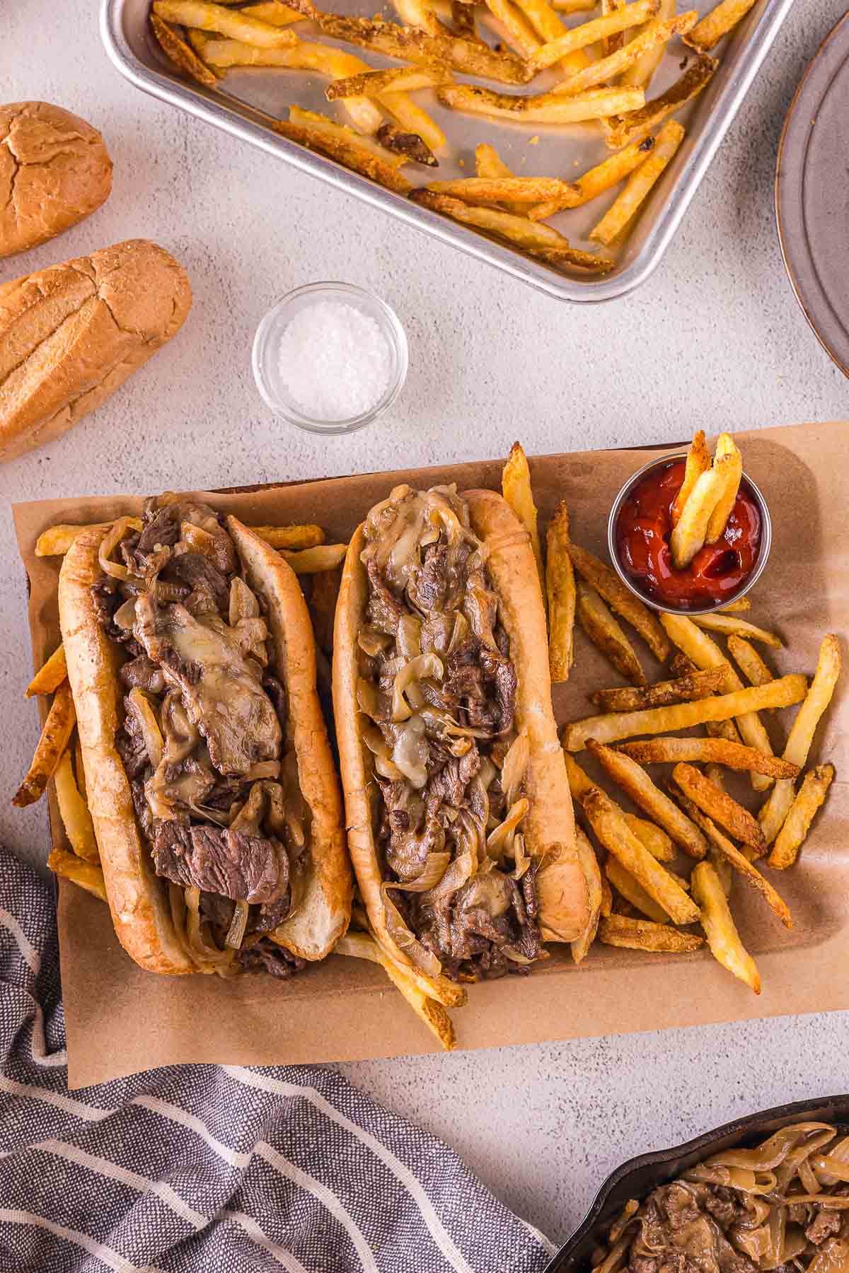 Two cheesesteak sandwiches with sautéed onions on a tray, served with French fries, a small cup of ketchup, and a dish of salt.
