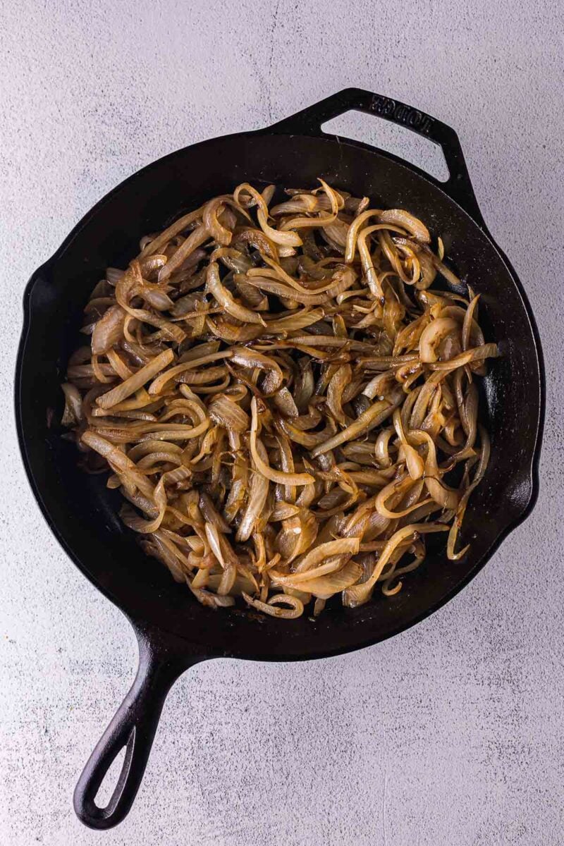 Sliced onions being caramelized in a black cast iron skillet on a light-colored surface.