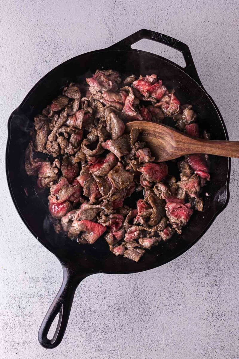 Sliced beef cooking in a black cast iron skillet, being stirred with a wooden spoon on a light-colored surface.