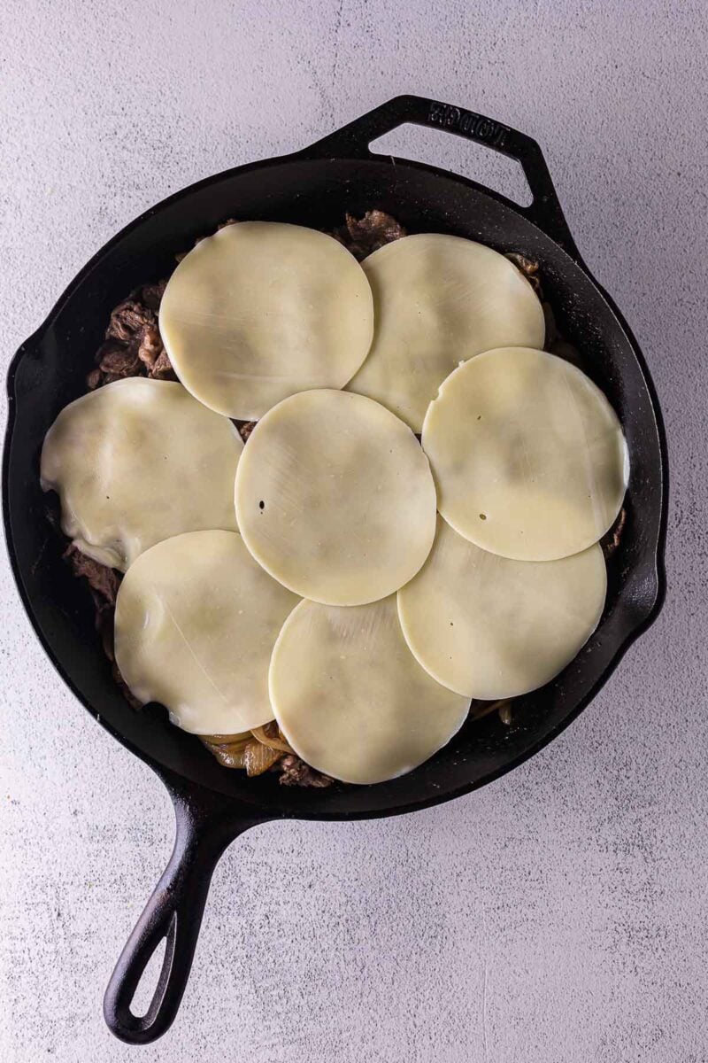 A cast iron skillet filled with cooked meat and vegetables, topped with overlapping slices of provolone cheese.