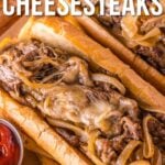 Two Philly cheesesteak sandwiches with melted cheese, sautéed onions, and sliced beef in hoagie rolls, served on a wooden platter with a side of ketchup.