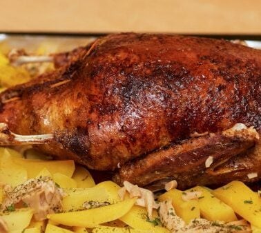 A whole roasted duck is placed on a tray surrounded by sliced potatoes and herbs.