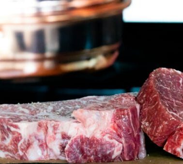 Raw marbled beef steaks and chunks are placed on a cutting board with a copper pot in the blurred background.