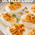 A plate of deviled eggs topped with a spicy, orange filling and garnished with herbs and bacon bits. Text on image reads "How to make spicy deviled eggs.