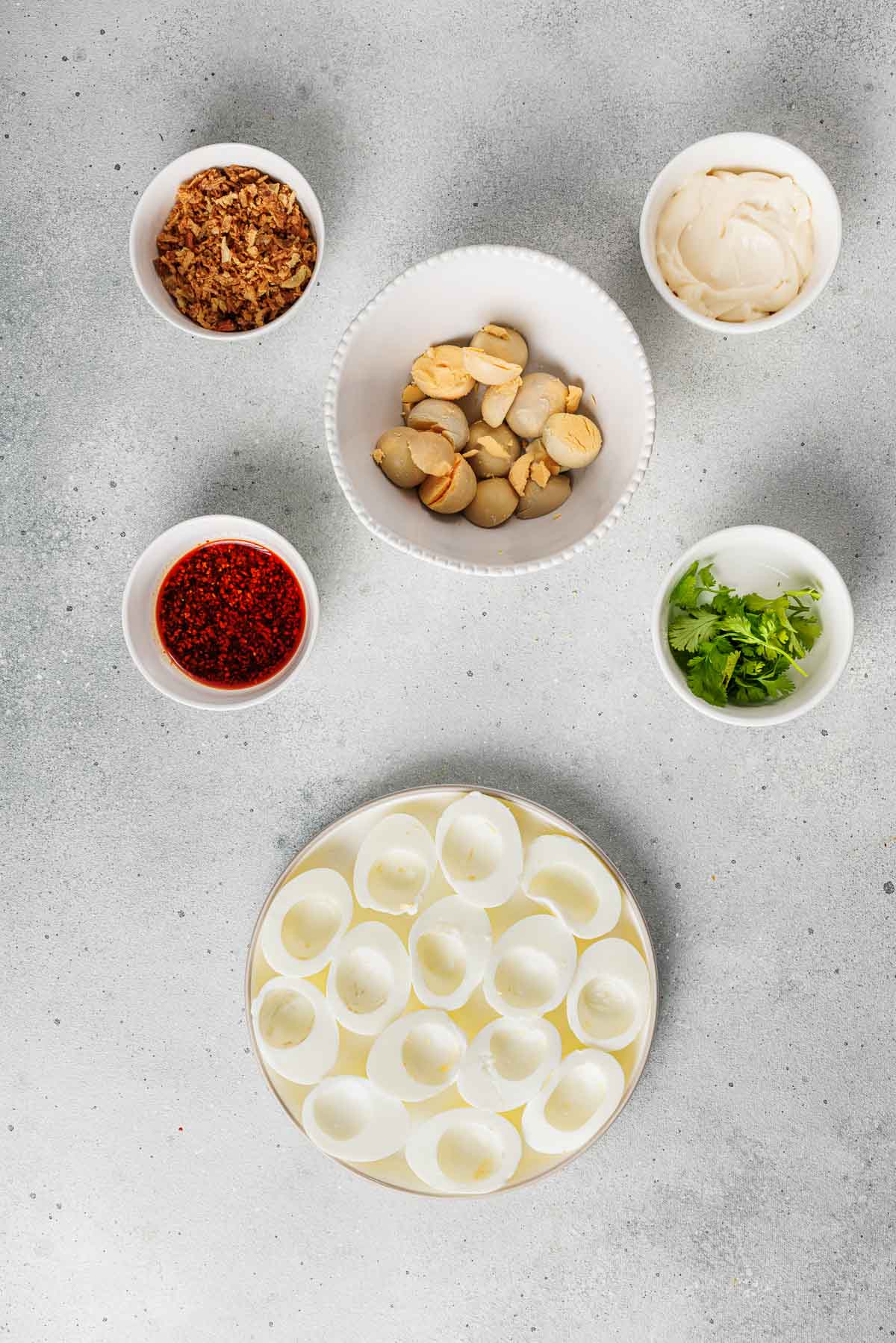 Bowls containing fried shallots, quail eggs, mayonnaise, chopped cilantro, chili sauce, and empty egg white halves arranged on a gray surface.