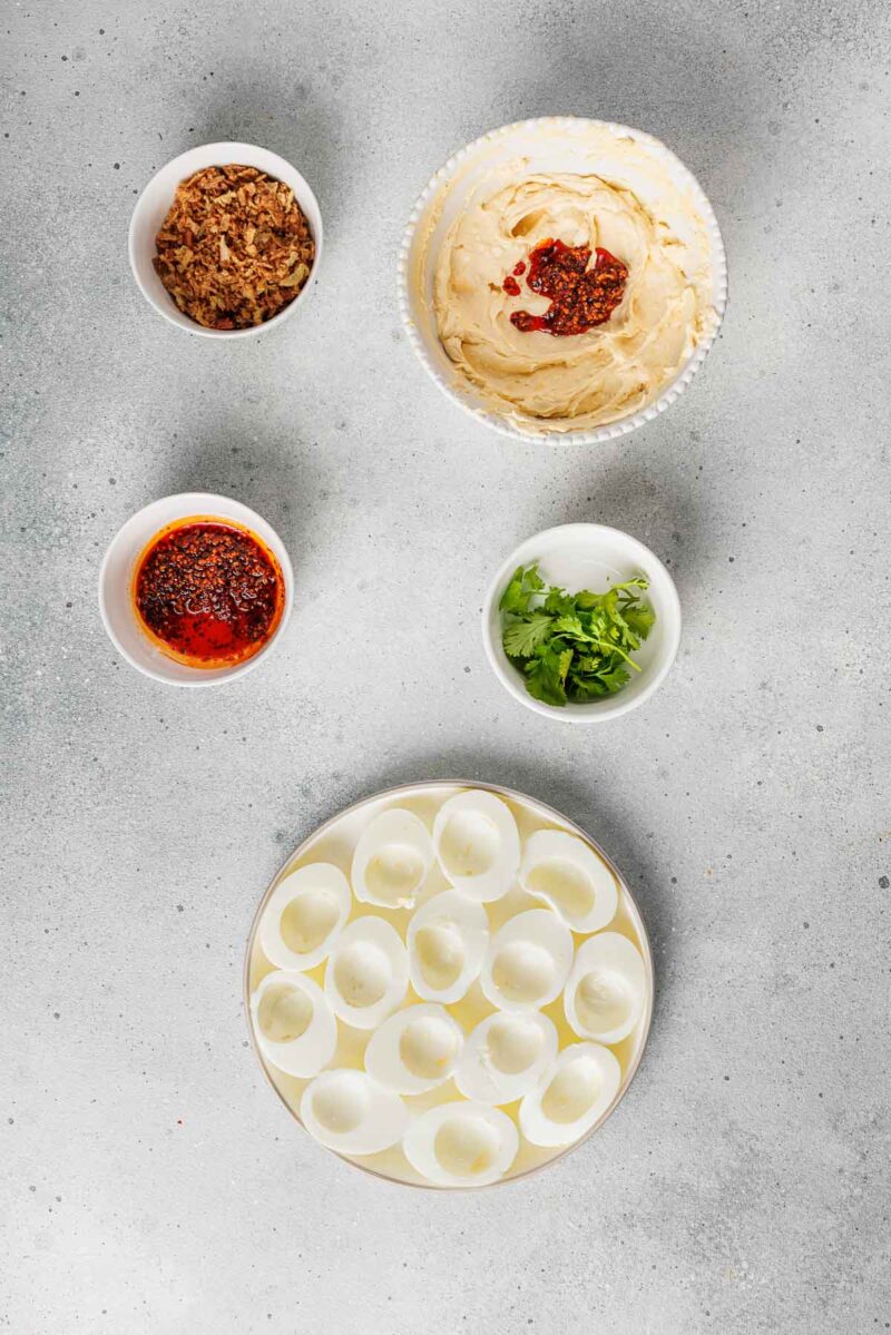 A plate of empty deviled egg whites is surrounded by bowls of hummus with chili oil, crispy onions, chili oil, and chopped cilantro on a gray surface.