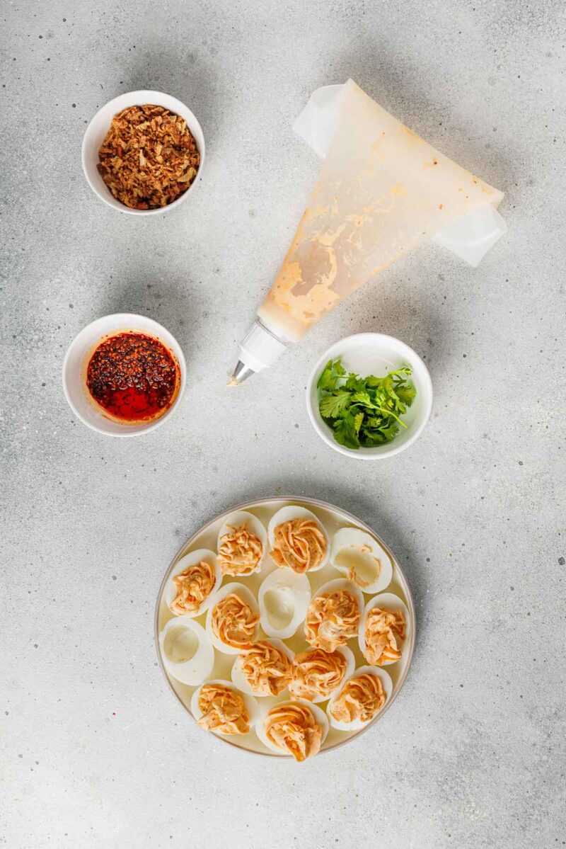A plate of deviled eggs with piped filling is surrounded by bowls of fried onions, chili oil, cilantro, and a piping bag on a gray surface.