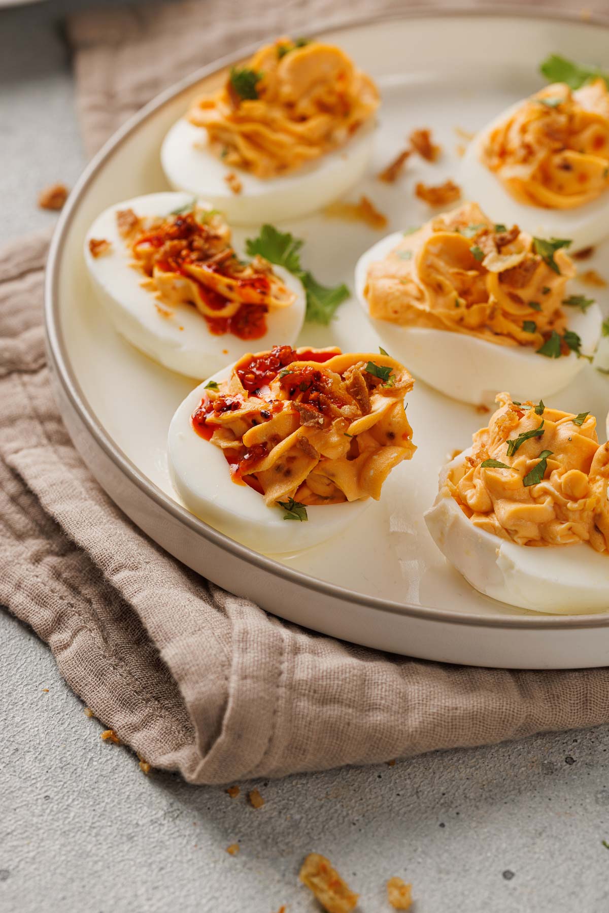 A plate of deviled eggs topped with herbs and crispy chili garlic sauce, arranged on a light-colored plate with a beige napkin underneath.