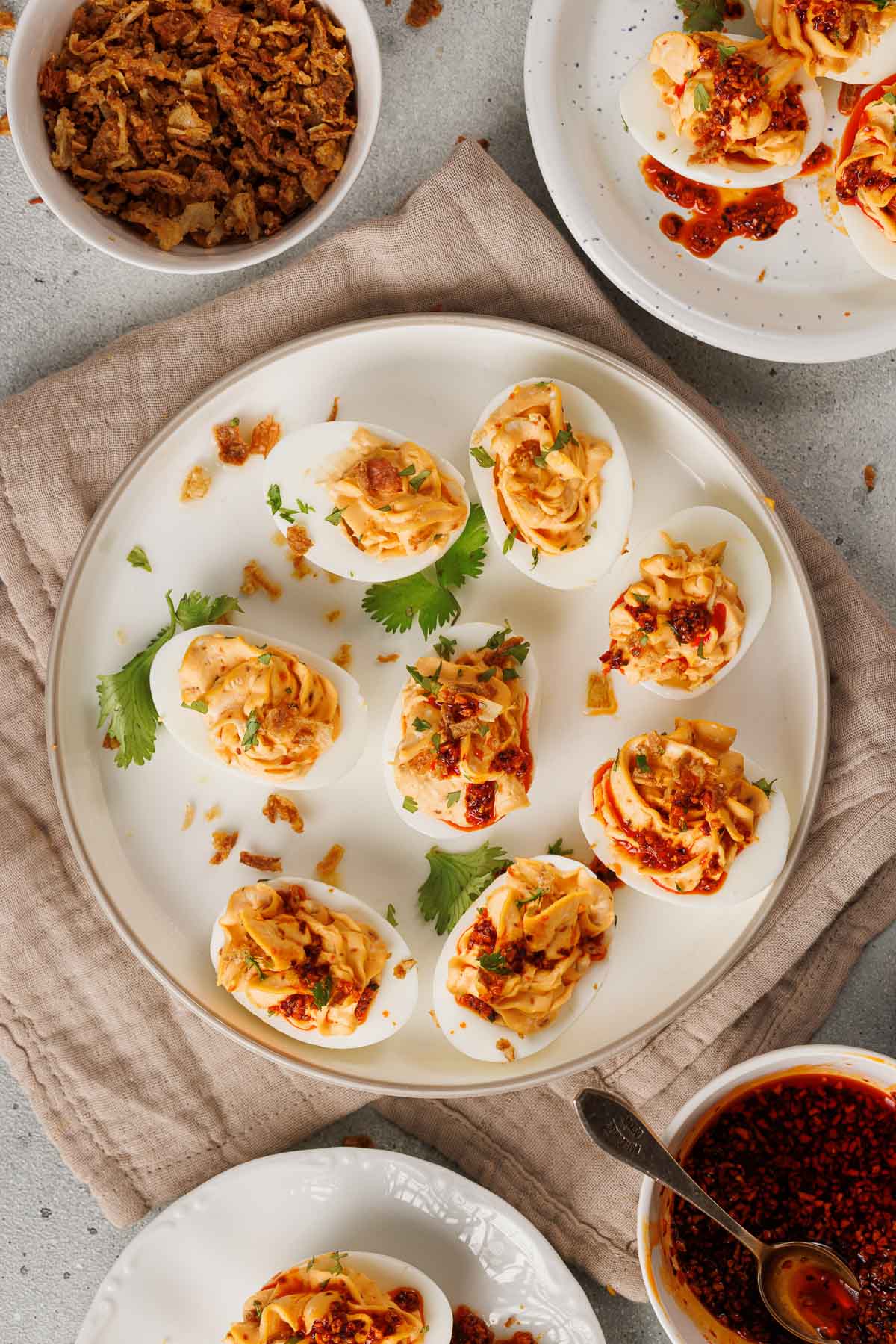 A plate of deviled eggs garnished with spicy chili garlic sauce, herbs, and crispy onions, with extra sauce and toppings in bowls nearby.