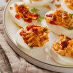 A plate of deviled eggs topped with a chili garlic sauce and herbs, arranged on a light round dish placed on a beige cloth.