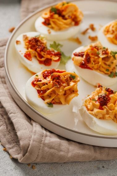 A plate of deviled eggs topped with a chili garlic sauce and herbs, arranged on a light round dish placed on a beige cloth.