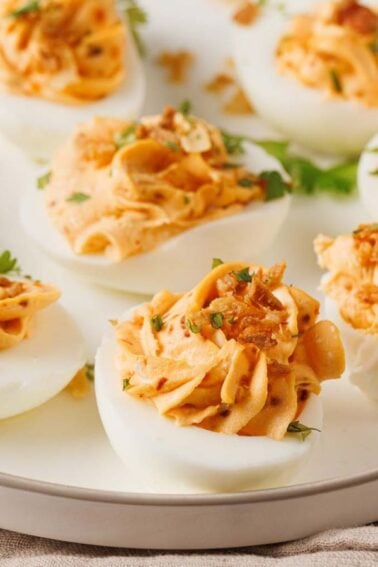 A white plate with eight spicy deviled eggs, each topped with a creamy orange filling, chopped herbs, and crunchy bits, on a beige cloth.