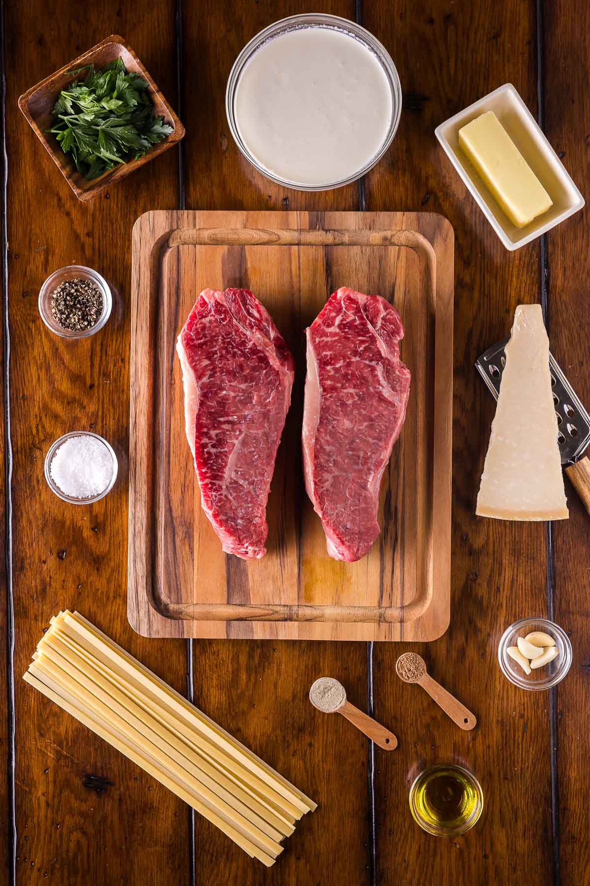 Raw steaks on a wooden cutting board surrounded by pasta, butter, cream, grated cheese, parsley, garlic, olive oil, salt, pepper, and spices on a wooden table.