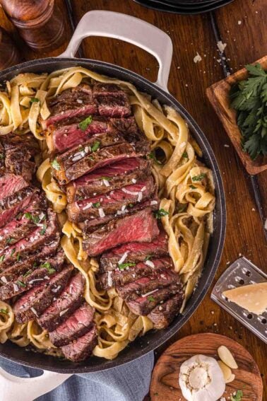 A skillet filled with sliced medium-rare steak arranged over fettuccine pasta, garnished with herbs and coarse salt. Nearby are a block of parmesan, garlic, and fresh parsley.